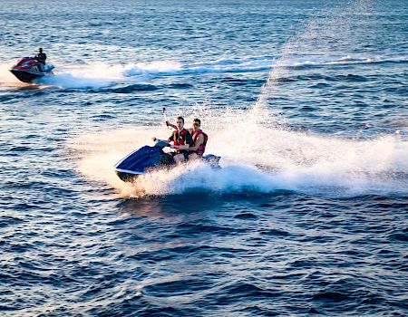 Two people ride a jet ski at high speed on choppy blue water, leaving white spray behind them, with another jet ski in the background.
