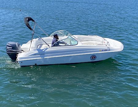 A small white motorboat with a black outboard sits on calm blue-green water; a person wears a cap in the cockpit near a windshield.