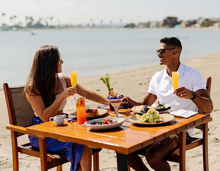 A couple sits at a wooden table on a sunny beach, enjoying drinks and a meal by the shore as the sea and palm trees stretch in the background.