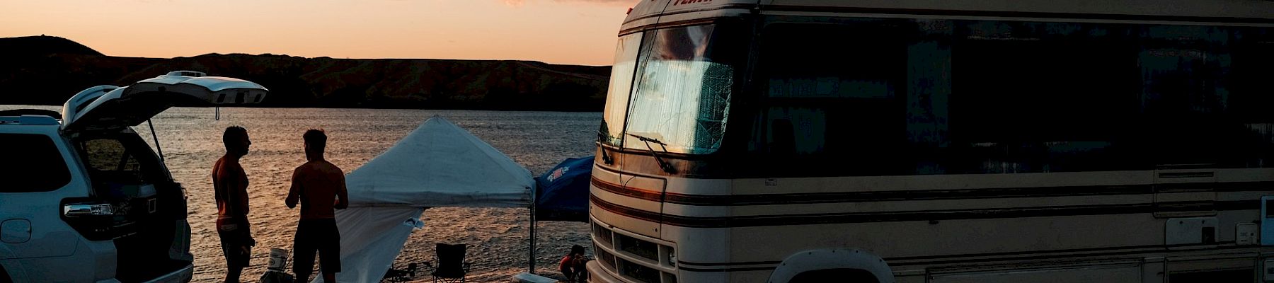 A coastal campsite at sunset with a parked RV, a tent, and a few people near a car, silhouettes against a orange-pink sky ending.