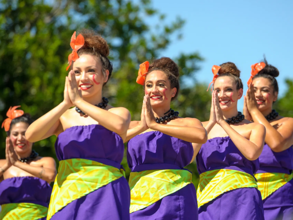 A group of five women in matching purple dresses with yellow-green sashes, orange bows, and red lipstick dance with hands pressed together, outdoors.