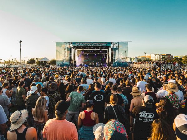 A large outdoor concert or festival with a big crowd gathered in front of a stage under clear skies, enjoying the performance.