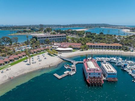 Aerial view of a coastal marina with docks, boats, and a sandy beach, plus resort buildings and clear blue water, all under sunny skies.