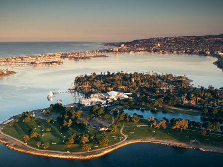 Aerial view of a coastal island park with green lawns, trees, water channels, and a small settlement along the shore at sunset.