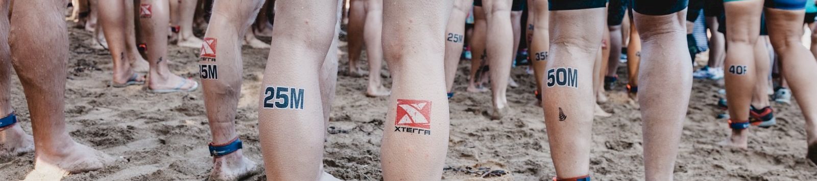 A crowd of runners standing on a sandy beach, wearing numbered ankle bands and athletic shorts, ready for a race.