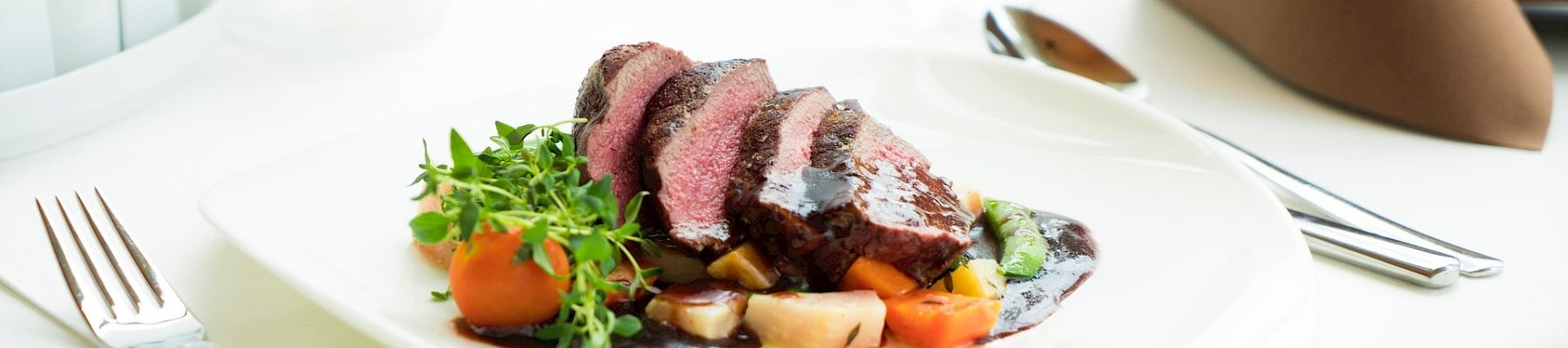 A plated steak with vegetables and greens on a white table, set with silverware and a fancy napkin, in a bright dining scene.