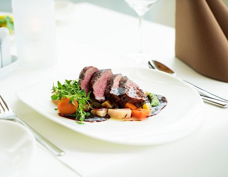 A plated steak with vegetables and greens on a white table, set with silverware and a fancy napkin, in a bright dining scene.