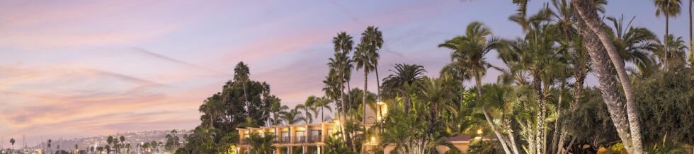 A tranquil beach at sunset with calm waves, palm trees, and cozy cabanas along the shore, glowing pastel sky reflecting on the water.