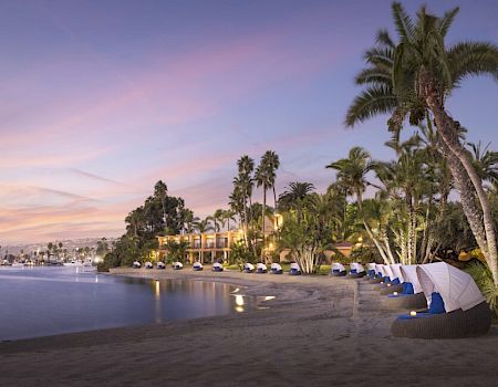 A tranquil beach at sunset with calm waves, palm trees, and cozy cabanas along the shore, glowing pastel sky reflecting on the water.