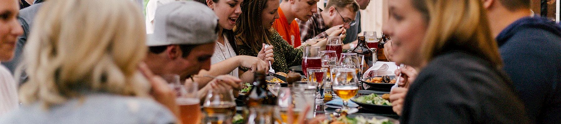 A long table of people dining together outdoors, sharing food and drinks at a social meal.
