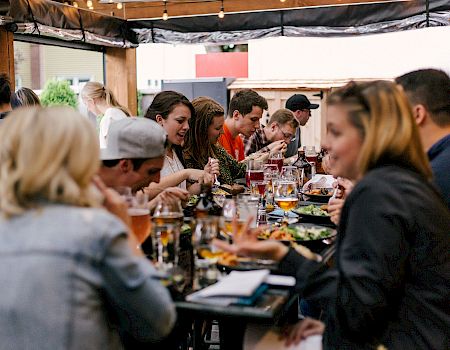 A long table of people dining together outdoors, sharing food and drinks at a social meal.