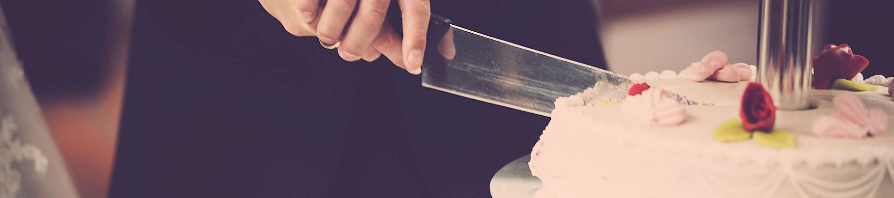 A person cuts a wedding cake with a knife, a tiered cake decorated with flowers in the foreground.