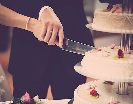 A person cuts a wedding cake with a knife, a tiered cake decorated with flowers in the foreground.