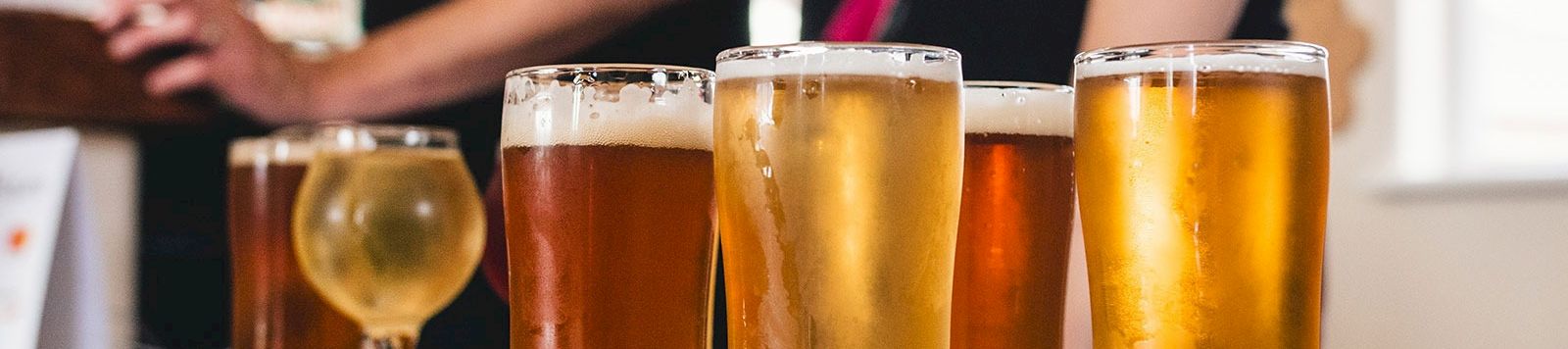 Three pints of beer on a wooden flight board with two bartenders in the background, indoors, casual bar scene.