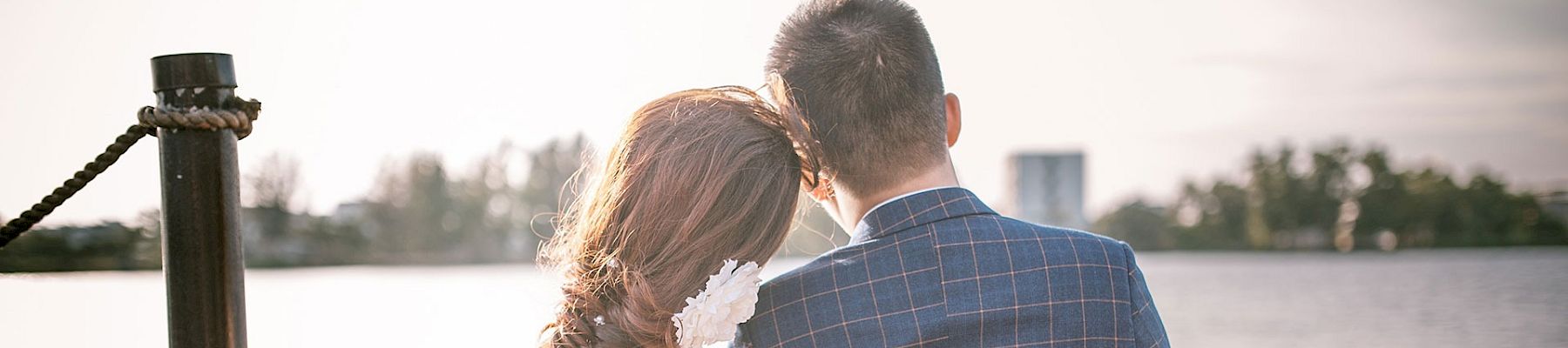 Two people sit close on a dock by the water, wearing a wedding dress and suit, sharing a quiet, intimate moment by the lake.
