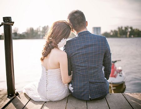 Two people sit close on a dock by the water, wearing a wedding dress and suit, sharing a quiet, intimate moment by the lake.