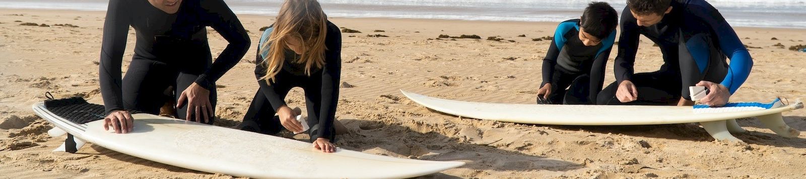 Four people on a sunny beach prepare two surfboards, kneeling on the sand while checking their plans near the ocean.
