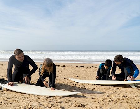 Four people on a sunny beach prepare two surfboards, kneeling on the sand while checking their plans near the ocean.