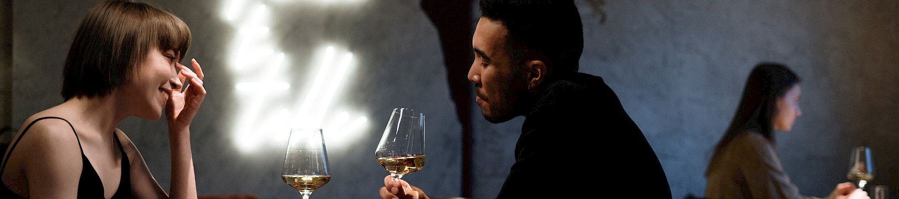 Two people sit per table in a dim, moody restaurant; a man and woman share a wine moment while others blur in the background, lights glow softly.