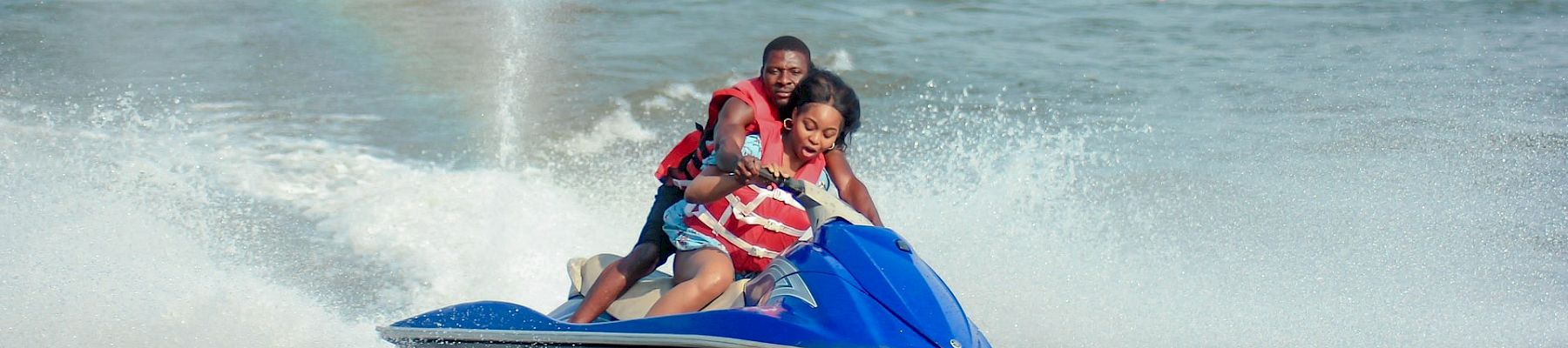 A couple rides a blue jet ski fast across a lake, creating a spray as they hold on tightly and smile, with a distant shoreline in view.