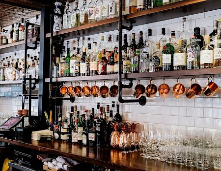 A well-stocked bar with bottles on shelves, copper mugs, and row of hanging glasses behind a polished wooden counter, ready for service.