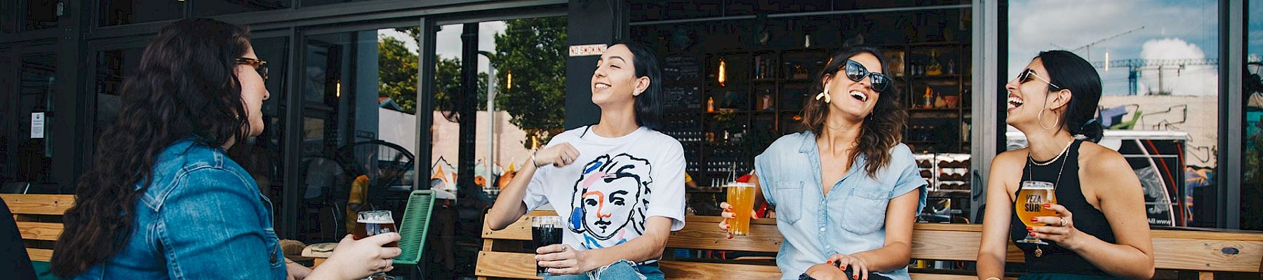 A group of friends sits at an outdoor cafe, chatting and enjoying drinks around a wooden table by a glass-front bar.