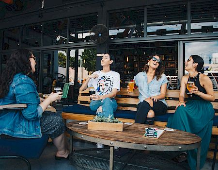 A group of friends sits at an outdoor cafe, chatting and enjoying drinks around a wooden table by a glass-front bar.