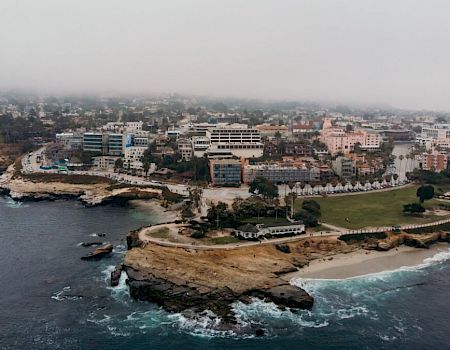 Coastal city view with rocky shoreline, waves crashing against cliffs, and mid-rise buildings along the coast, under a hazy sky.