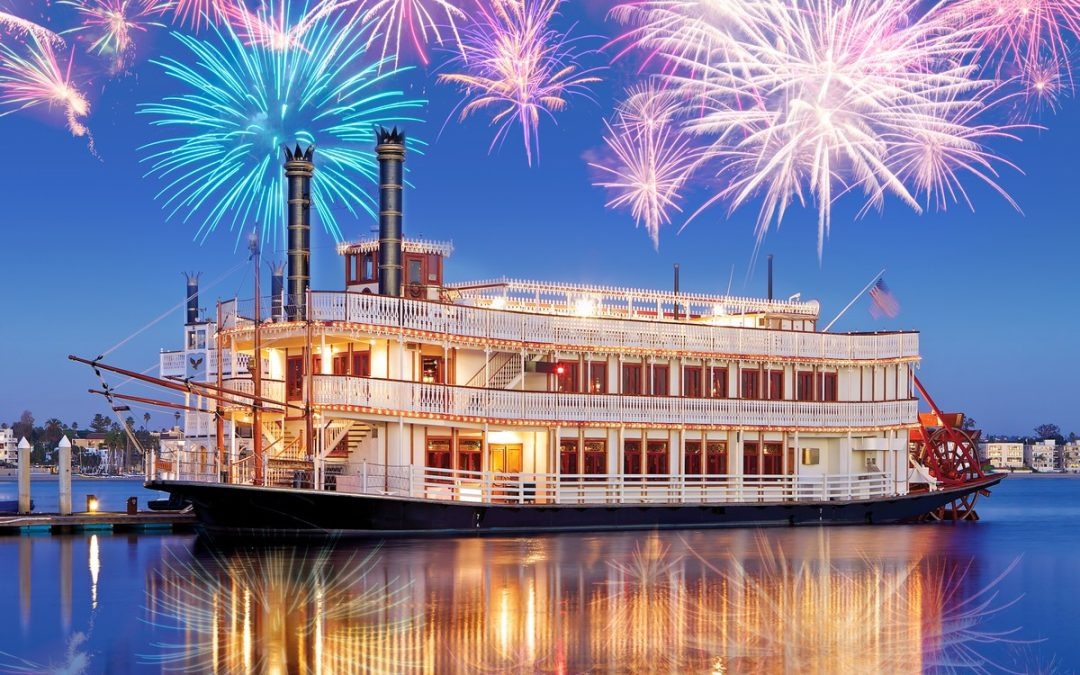 A large, multi-deck riverboat on calm water with colorful fireworks overhead, reflecting in the water at dusk.