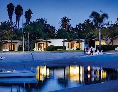 A serene seaside resort at dusk with palm trees, lit bungalow rooms along the shore, and calm water reflecting lights, a small boat docked nearby.