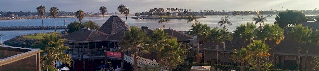 A tropical resort pool area with palm trees, lounge chairs, a thatched-roof bar, and a sandy beach beyond, under a partly cloudy sky.