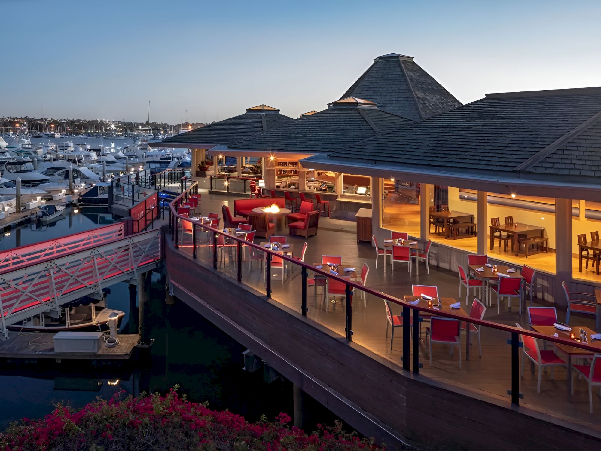 A waterfront restaurant on a deck over the water, with red chairs, warm lighting, and boats docked nearby.