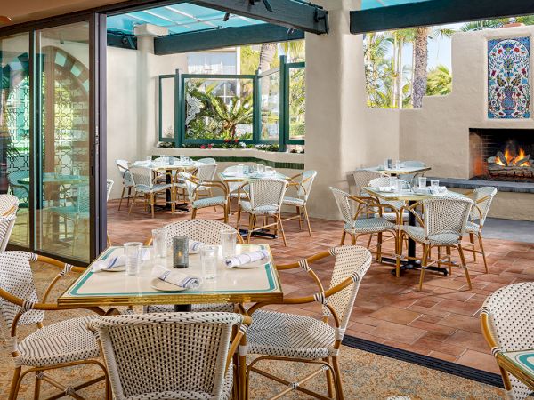 A bright, stylish outdoor dining area with wicker chairs, tables set with plates, and a fireplace in the wall.