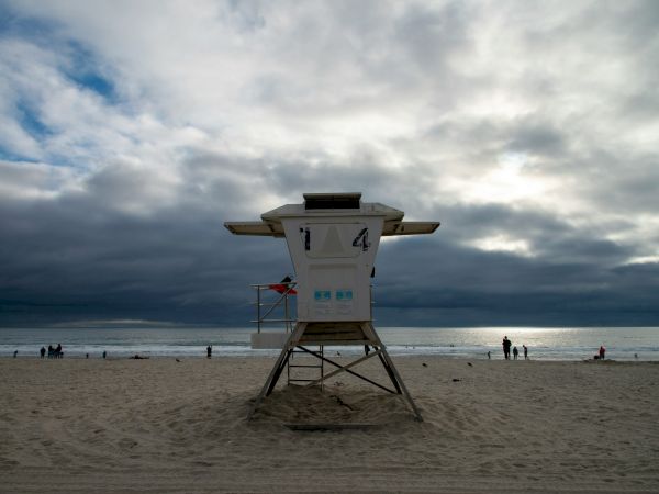 A lifeguard tower on a sandy beach with dark clouds over the ocean and a few distant beachgoers, ready to watch the waves.