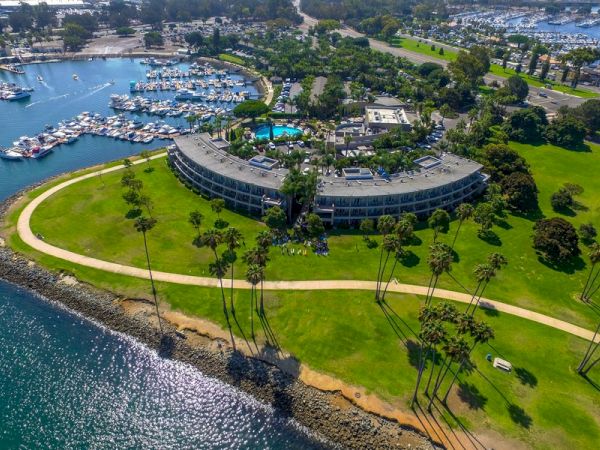 An aerial view of a curved, coastal hotel or resort building surrounded by green lawns, palm trees, a winding path, and a marina nearby, with water along the shoreline.