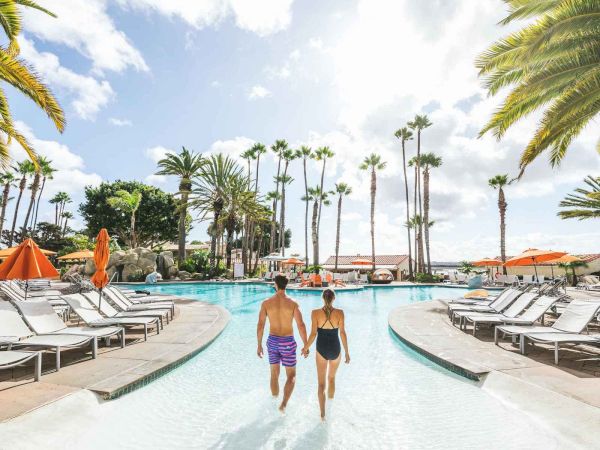 Two people walk hand-in-hand through a resort pool, surrounded by palm trees, lounge chairs, and sunny skies, enjoying a tropical vacation.