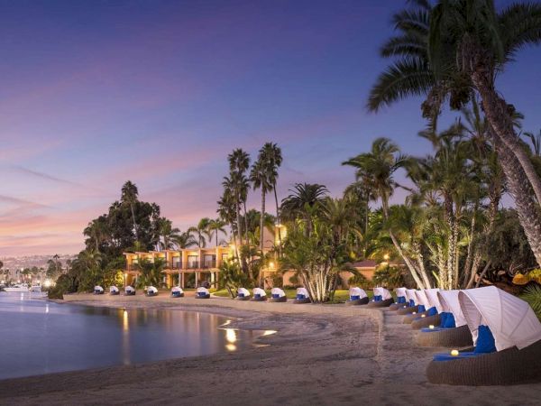A tropical beach at dusk with calm waters, palm trees, and line of blue-and-white cabanas along the shore, reflecting a tranquil resort vibe.