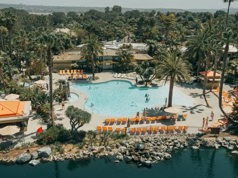 A resort pool area with palm trees, sun loungers, orange umbrellas, and rocky shoreline along clear blue water.