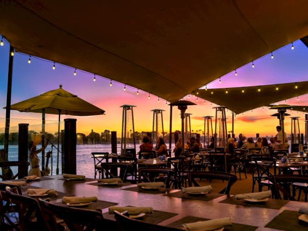 A lively waterfront restaurant at sunset with string lights, umbrellas, and people dining under a canopy by the water.