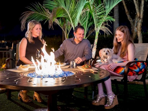A family sits around a glowing fire pit on a patio at night, smiling and chatting, with a small dog on a woman's lap.