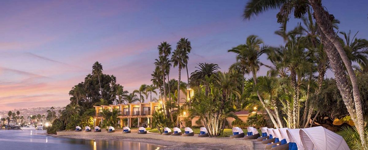 A tropical resort beach at twilight, palm trees, calm water, luxury huts lined along the shore with soft lighting.