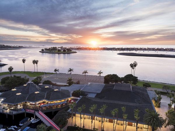 Coastal sunset over a waterfront resort with palm trees, a boardwalk, and calm water, buildings lighting up as the sun dips below the horizon.