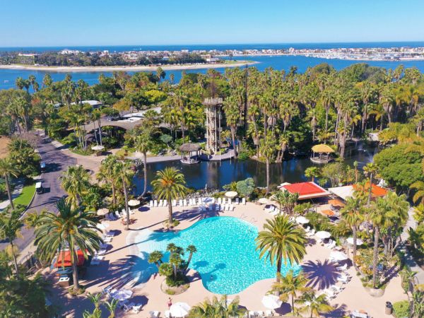 A tropical resort with a large blue pool, palm trees, sunlit lounging areas, and water channels or a lagoon in the background.