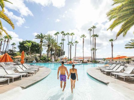 Two people walk hand‑in‑hand through a resort pool, palm trees frame the scene, loungers and orange umbrellas line the edges under a sunny sky.