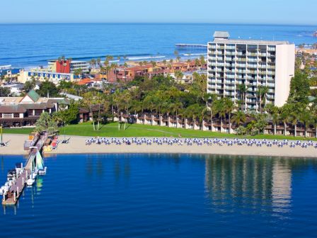 A coastal resort with a sandy beach, palm trees, high-rise hotel, and a calm blue sea; a pier extends into the water near a treeline.
