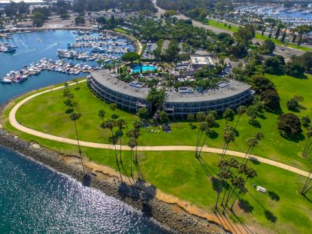 Aerial view of a curved seaside resort with a circular building, green lawns, palm trees, a winding path along the water, and a marina nearby.