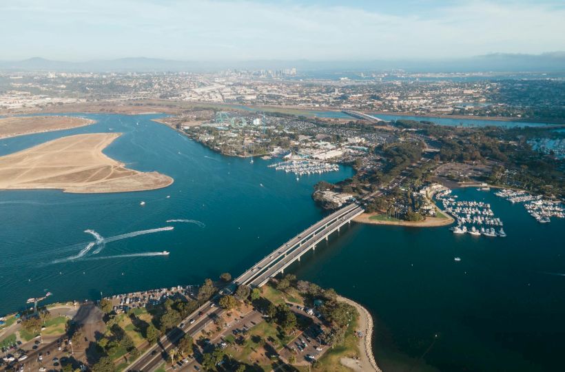 Aerial view of a coastal city with a winding river, sandy barrier beach, marina, and a bridge connecting landmasses, sunny and calm water.