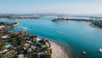 A scenic coastal view of a tropical beach with palm trees, clear turquoise water, boats, and a distant bridge across the bay, sunny day.