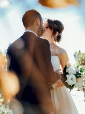 A couple in wedding attire share a kiss, framed by soft, warm bokeh and a bouquet of white flowers, celebrating love in a sunny outdoor setting.