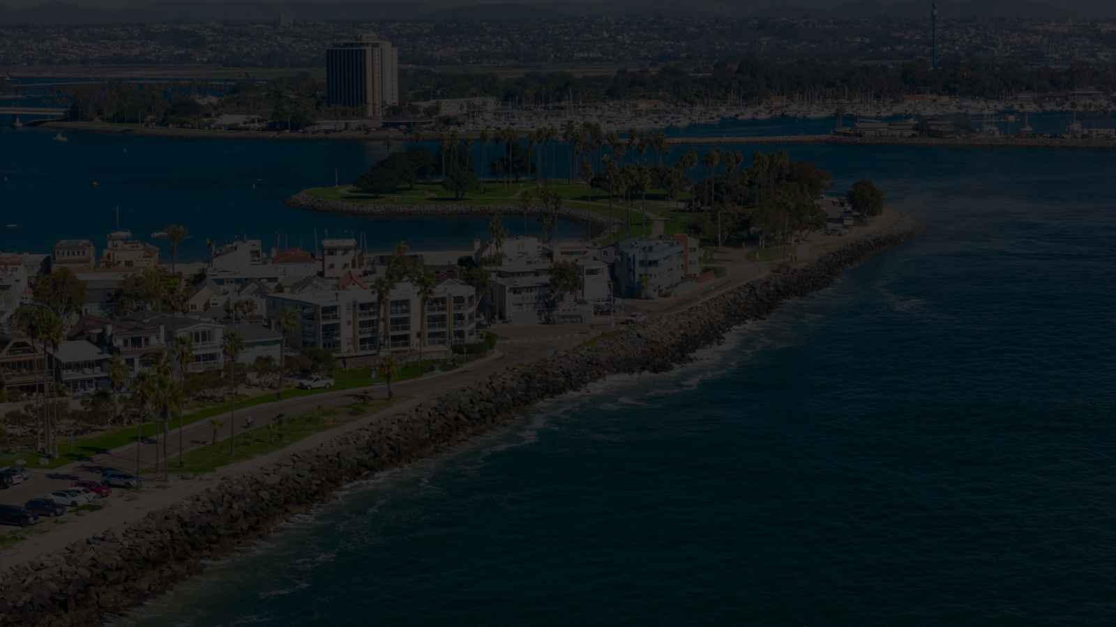 Aerial view of a coastal town with houses along a narrow strip of land, blue water on one side and a rocky shoreline on the other, calm and sunny.